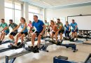 Groupe mixte de sportifs réalisant un relais sur des rameurs dans une salle lumineuse, avec un panneau de collecte solidaire sans inscription visible.