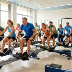 Groupe mixte de sportifs réalisant un relais sur des rameurs dans une salle lumineuse, avec un panneau de collecte solidaire sans inscription visible.